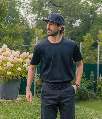 Abgebildet ist ein Mann, der ein schwarzes Mercedes-Benz Basic T-Shirt mit Sternlogo und eine schwarze Mercedes-Benz Cap trägt.