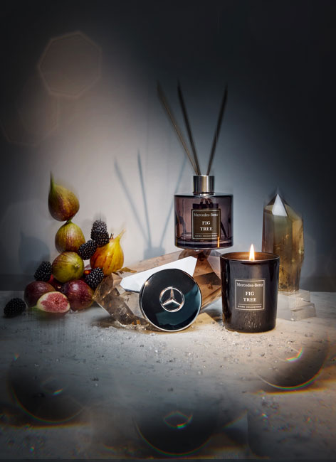 Abgebildet wird die Mercedes-Benz Raumduft Collection mit Kerze und Diffuser. Die Artikel sind in einem dunkleren Set dargestellt mit Lichteffekten und Kristallen sowie Feigen als Dekoelemente.