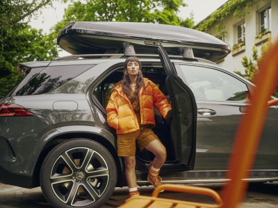 Abgebildet ist eine junge Frau in orangener Jacke. Sie steht in der geöffneten Hintertür eine grauen Mercedes-Benz, welcher eine Mercedes-Benz Dachbox montiert hat.