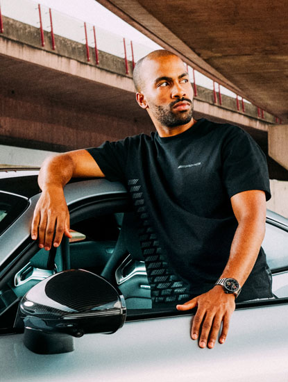 Zu sehen ist ein junger Mann in schwarzem AMG Logoshirt, der an einem silbernen Mercedes AMG lehnt und zur Seite schaut.