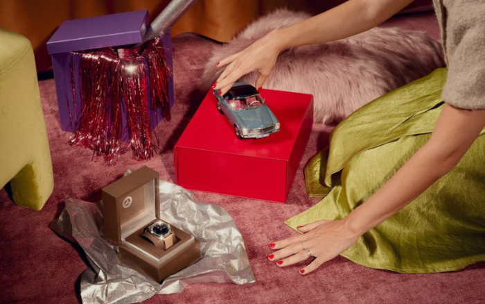 Zu sehen ist eine Geschenkeszene auf einem rosa Teppich.eine Person stellt ein Modellauto auf eine rote Geschenkbox. weitere Geschenkideen sind in Boxen zu sehen, darunter eine Armbanduhr.