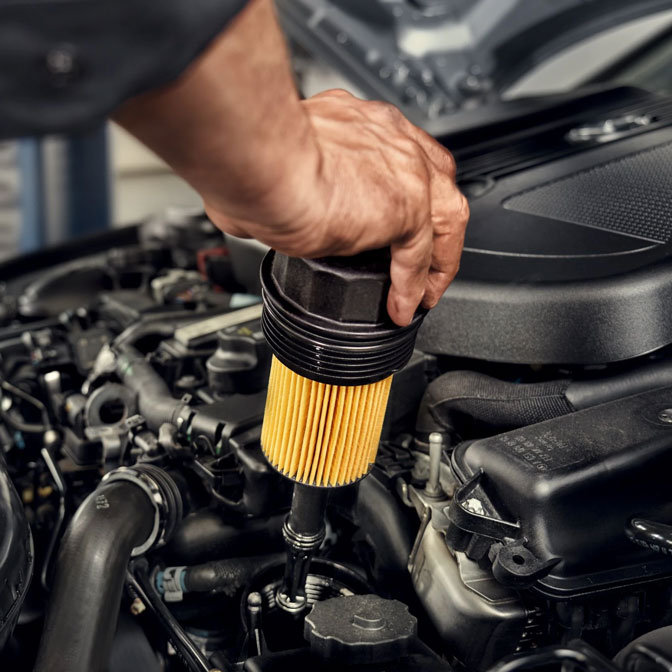 Gezeigt wird ein original Mercedes-Benz Ölfilter, der gerade in den Motorraum eingebaut wird.