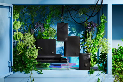 Mercedes-Benz Collection Sommer Kampagne Zu sehen sind verschiedene schwarze Lederwaren der Mercedes-Benz Collection. Die Brieftaschen sind vor einem bunten Hintergrund präsentiert - in einem hellblauen Regal mit grünen Pflanzen dekoriert.