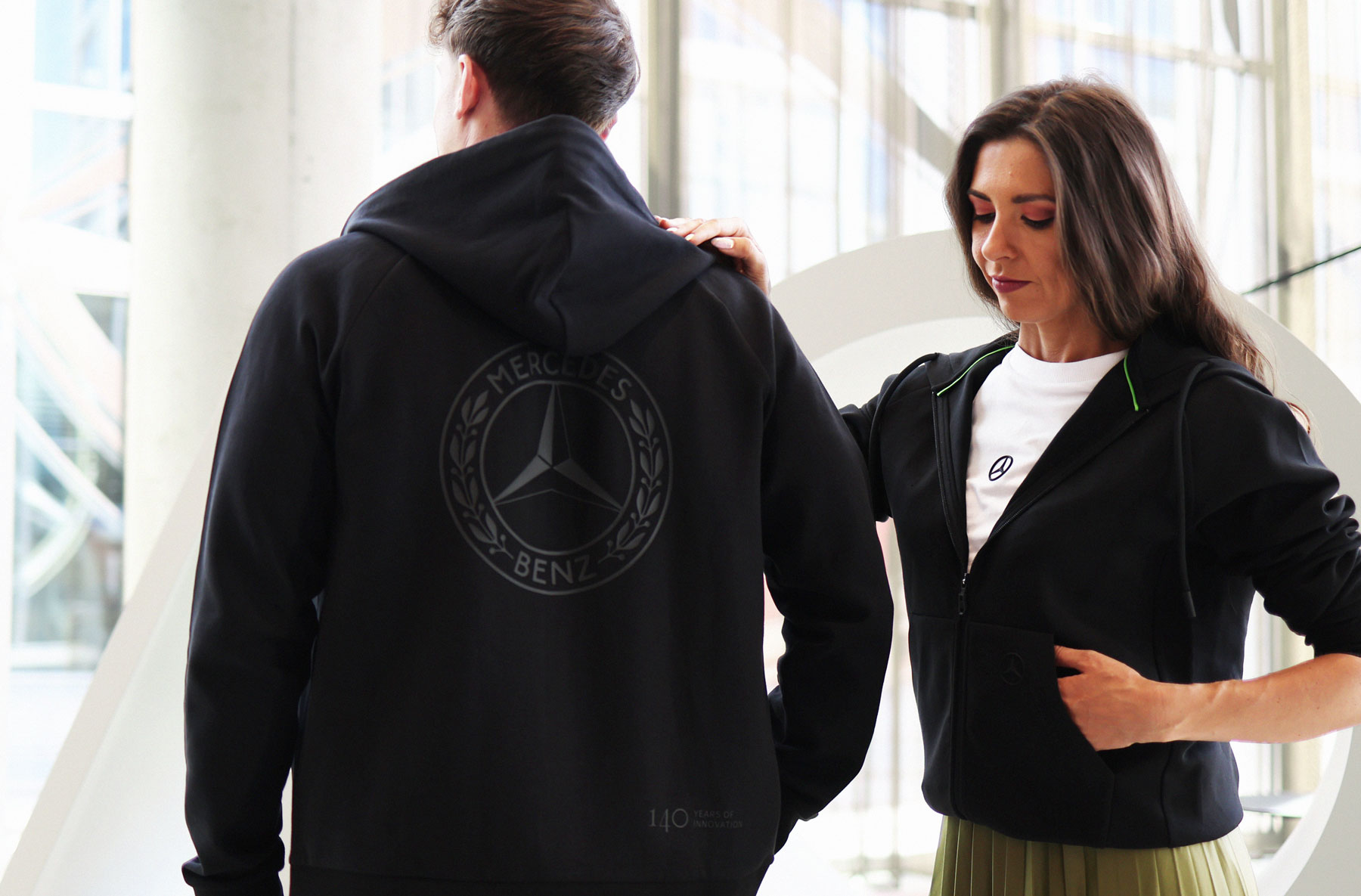 Schwarze Sweatshirt-Jacke der Mercedes-Benz „140 Years of Innovation Collection“. Das Kleidungsstück wird von zwei Personen präsentiert, die Rückseite zeigt ein großes Mercedes-Benz Logo.